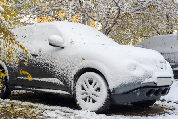 A gray car covered with the first snow against the background of yellow trees. First snowfall. Problems for drivers in winter. Bad weather conditions. Change of weather.