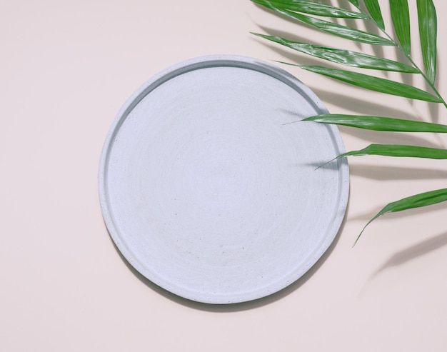 Gray empty decorative plate and green palm leaf on a beige background product and cosmetic display stage top view