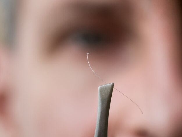 Gray hair on tweezers on blurred face.