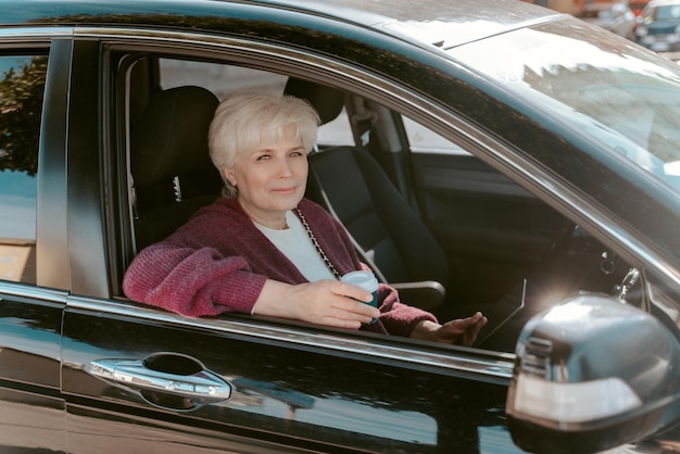 Gray-haired lady seated in the motor vehicle with a tablet computer and a disposable coffee cup