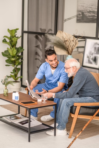 Photo gray-haired man seated at the coffee table beside his caregiver pointing with a ball-point pen at the newspaper article