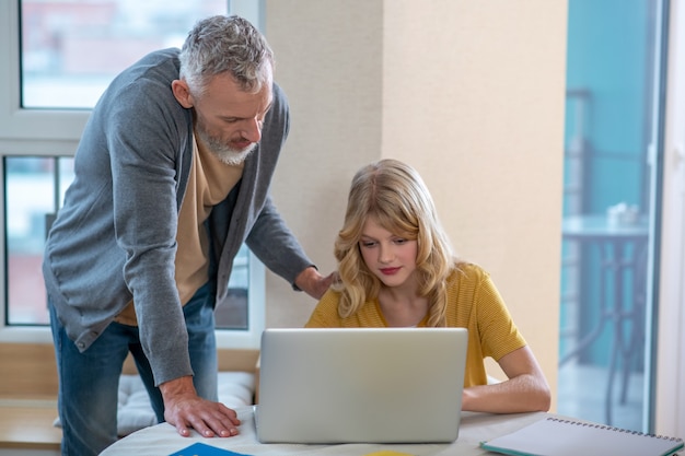 A gray-haired man standing next to his daughter while she working on the laptop