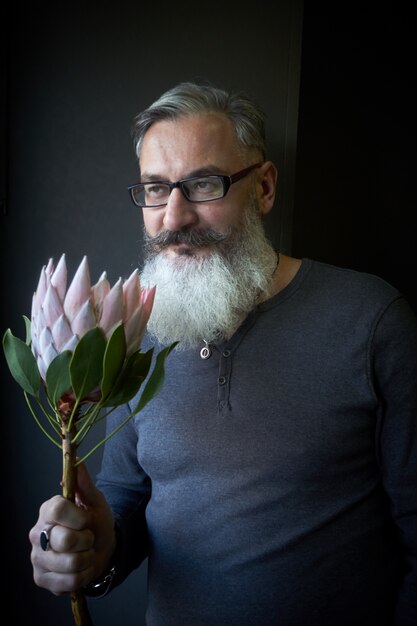 Gray-haired man with glasses and a beard holds a pink protea in his hands