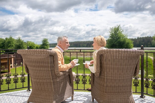 Gray-haired mature man and a smiling blonde lady sitting in the wicker arm-chairs with cups and saucers in their hands