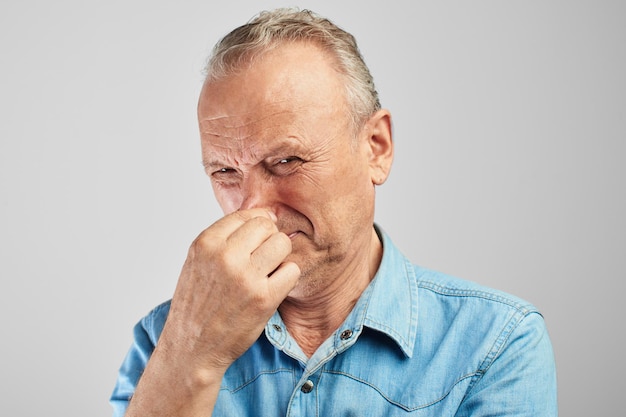 Gray-haired old man closes his nose with hand, feels disgusting smell of sweat, stinky feet and armpits against white wall