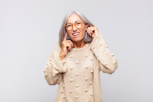 회색 머리의 여성은 화가 나고 스트레스를 받고 짜증이 나서 귀를 듣지 못하는 소음, 소리 또는 시끄러운 음악에 양쪽 귀를 가리고 있습니다.