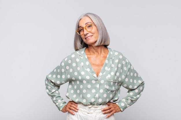 Gray haired woman looking happy, cheerful and confident, smiling proudly and looking to side with both hands on hips