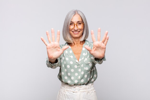 Gray haired woman smiling and looking friendly, showing number ten or tenth with hand forward, counting down