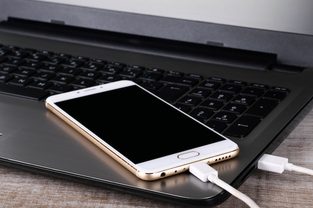 A gray laptop and a white telephone(smartphone) connected by a cable for data transmission on a wooden table. Data transfer. Information exchange