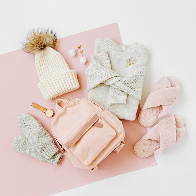 Gray warm knitted sweater with winter hat, mittens, backpack and fluffy fur slippers on pale pink background. Women's stylish winter clothes. Flat lay, top view.