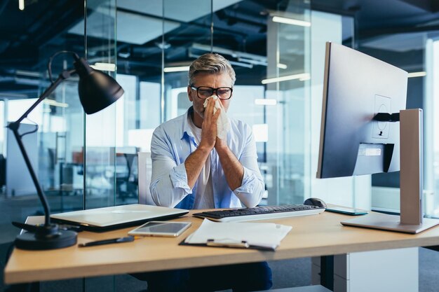 Grayhaired businessman with a runny nose working in the office at the computer cold and flu