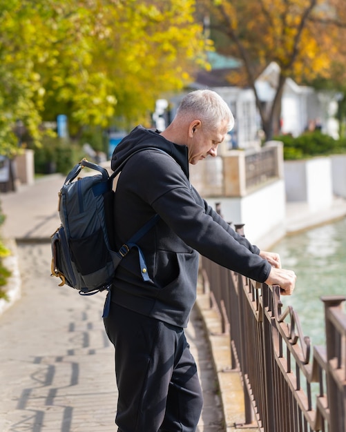 A grayhaired man with a backpack stands by a metal fence on the city embankment and looks at the water