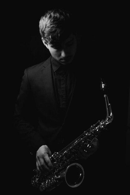 Grayscale shot of a cool and handsome guy holding his saxophone on a dark background