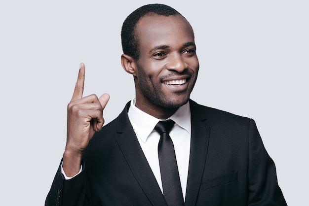 Great answer! Handsome young African man in formalwear pointing up and smiling while standing against grey background