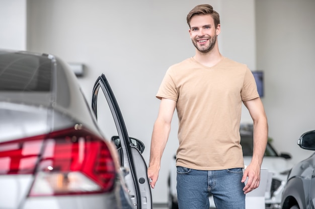 Great car. Happy attractive young adult man standing interested near new car in showroom opening front door