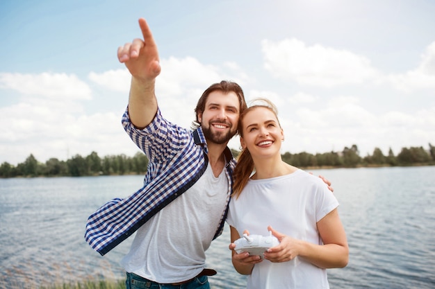 Great couple is standing together and looking up. Guy is hugging his wife and pointing up. Woman has control panel in hands. They look happy.