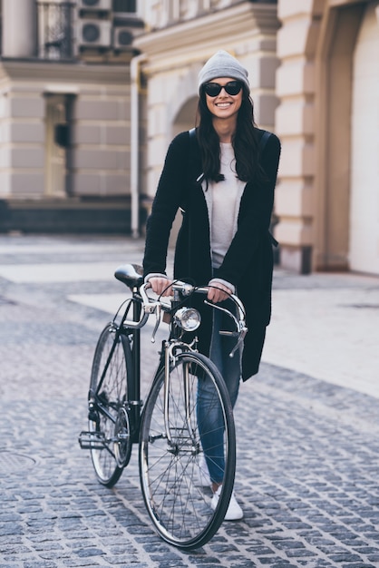 素晴らしい日。サングラスをかけた美しい若い女性が自転車を転がし、屋外を歩きながら笑顔でカメラを見て