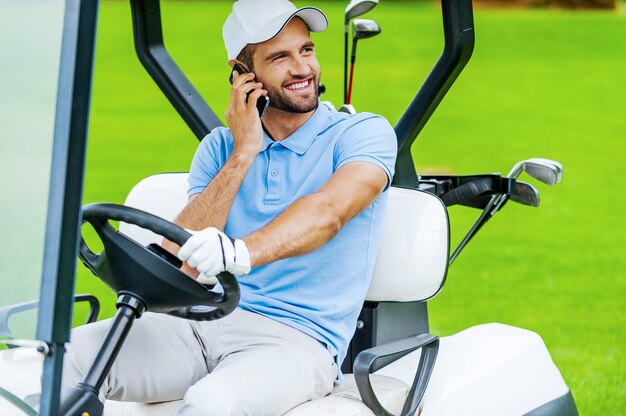 Great day to play golf! Handsome young smiling man driving a golf cart and talking on the mobile phone