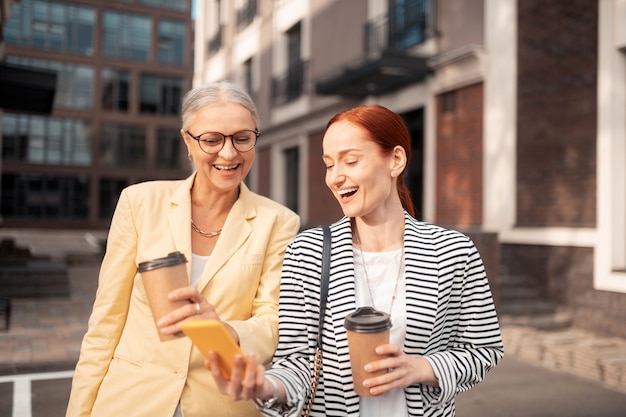 Gran giorno. due donne d'affari che ridono e guardano le foto sullo smartphone stando in piedi all'aperto