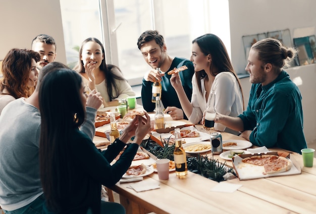 Great evening with friends. Group of young people in casual wear eating pizza and smiling while having a dinner party indoors