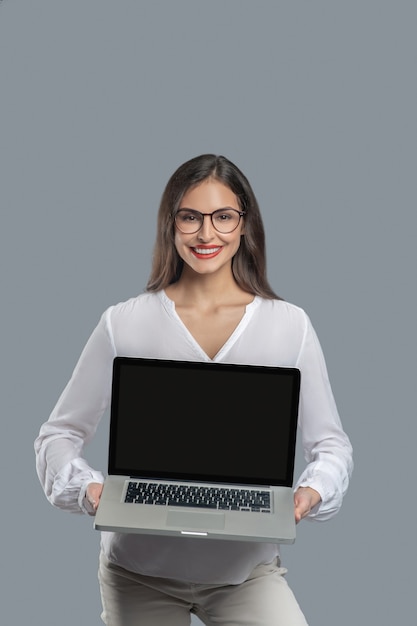 Great gadget. Joyful young long-haired woman with glasses holding open laptop in front of her showing screen