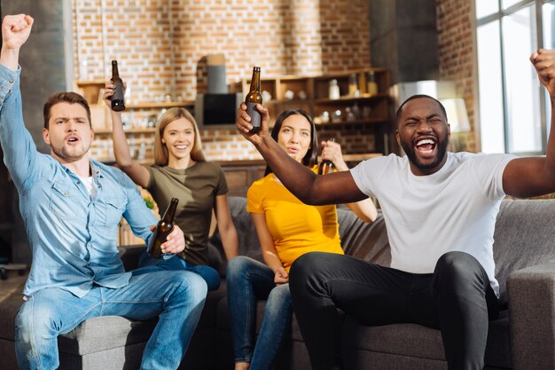 Great game. Four cute energetic excited friends drinking while sitting on the sofa and celebrating victory