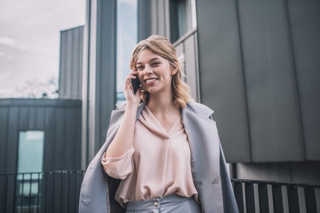 Great mood. Cheerful smiling young woman in gray jacket communicating on smartphone standing in afternoon outdoors near office