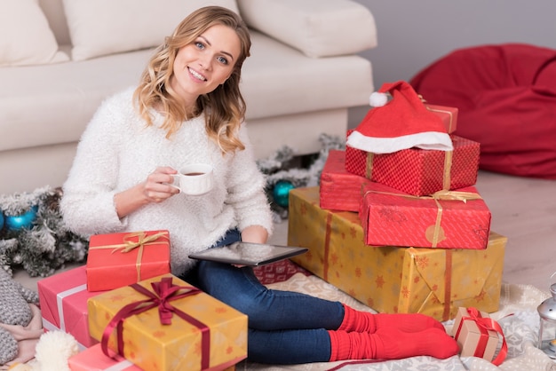 Great mood. Delighted attractive young woman sitting among Christmas presents and using her tablet while having tea