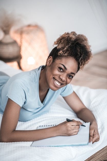 Great mood. Young adult black woman writing with pen in notebook lying on bed in bedroom in great mood