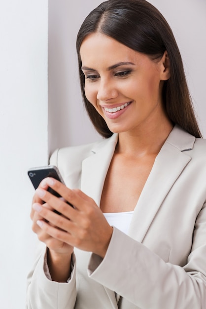 Great news. Beautiful young businesswoman in suit holding mobile and looking at it with smile