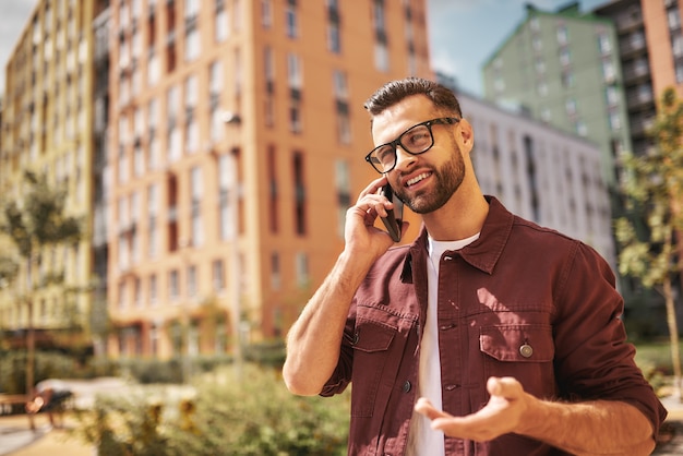 Great news Happy bearded man in casual wear and eyeglasses talking by phone and smiling while standing on the street. Lifestyle. Digital. Communication