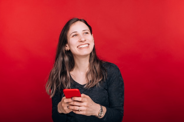 Great news. Smiling young woman looks up while she is holding her smart phone on red space.