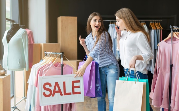 Great Offer. Two caucasian girlfriends are pointing at sale board, holding purchases, boutique interior, copy space