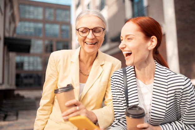 Great photos. two laughing businesswomen standing outdoors while holding paper cups of coffee and looking at photos on the smartphone