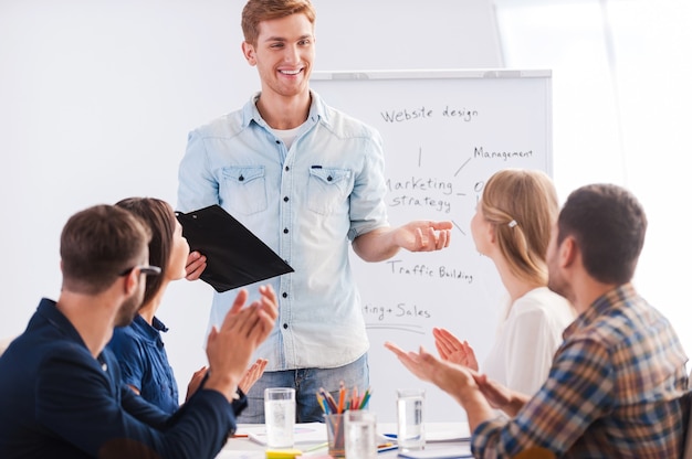 Great presentation! Group of business people in smart casual wear sitting together at the table and applauding to their colleague standing near whiteboard and smiling