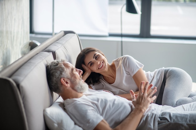 Great relationship. Bearded adult man telling smiling attentive woman listening lying on bed at home in afternoon