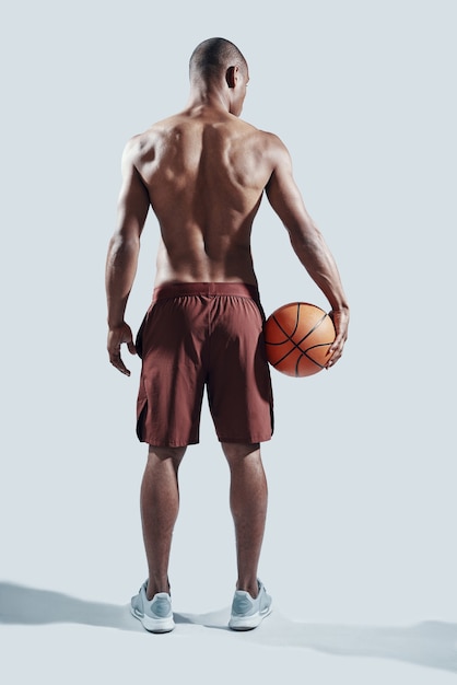 In a great shape. full length rear view of handsome young\
african man in sports clothing carrying basketball ball while\
standing against grey background