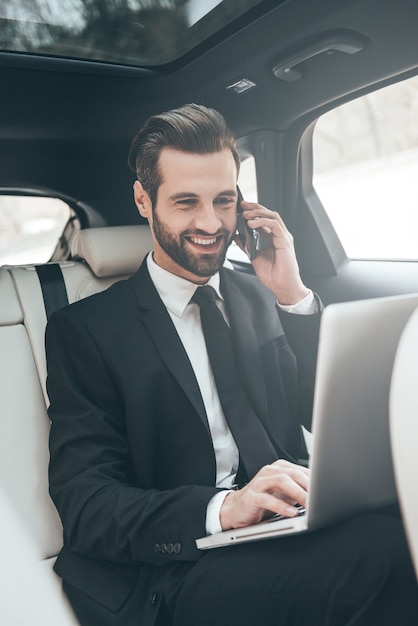 Great solutions every day. Handsome young businessman working on his laptop and talking on the phone while sitting in the car