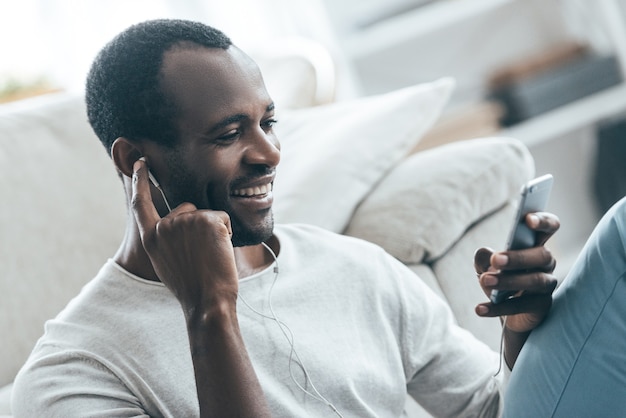 Bella canzone! bell'uomo africano che guarda il telefono cellulare e ascolta musica mentre è seduto sul tappeto a casa