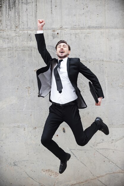 훌륭한 성공. 콘크리트 벽 앞에서 점프하는 동안 팔을 들고 긍정적인 표현을 하는 정장 차림의 행복한 청년
