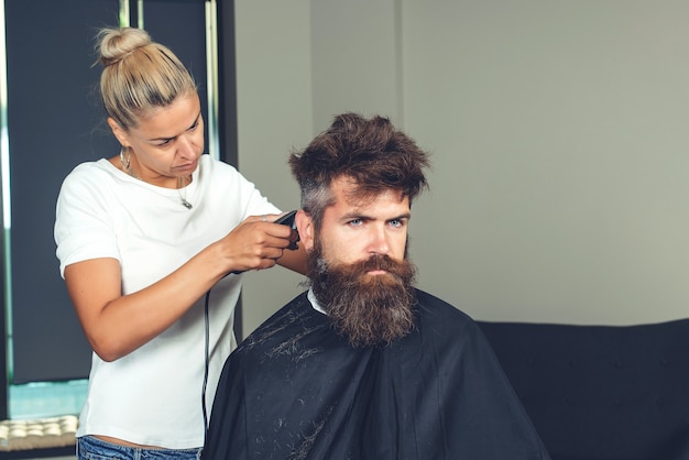 写真 理髪店での素晴らしい時間。理髪店の椅子に座っている間、美容師によって散髪されている陽気なひげを生やした男性。あごひげを生やしたハンサムな男が美容院で美容師に剃られている