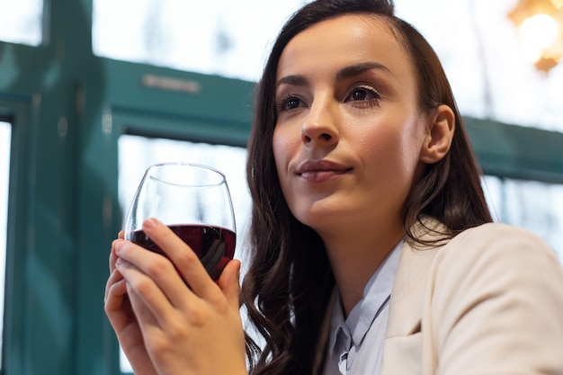 Great wine. Low angle of nice pleasant appealing woman holding glass of wine while posing near window and relaxing after work