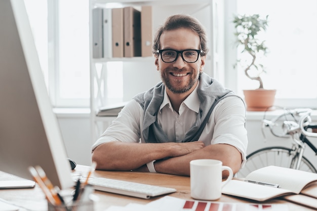 Great working day! Handsome young man looking at camera and smiling while sitting on working place in creative office