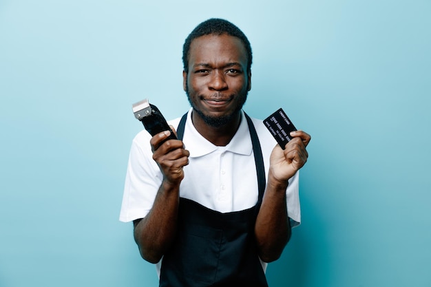 Greedy holding card with hair clippers young african american barber in uniform isolated on blue background