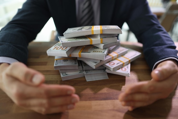 Greedy man in formal suit closes dollars pile with hands at desk in office businessman protects