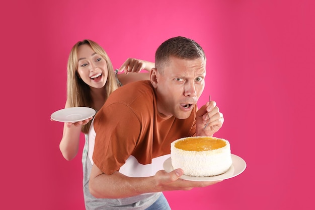 Greedy man hiding tasty cake from woman on pink background