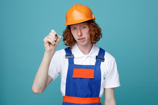 Greedy young builder man in uniform holding out marker pen to camera isolated on blue background