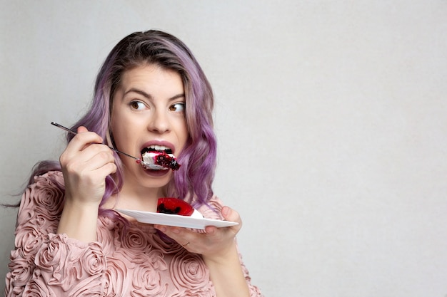 灰色の背景の上においしいチーズケーキを食べる紫色の髪の貪欲な若い女性。テキスト用のスペース