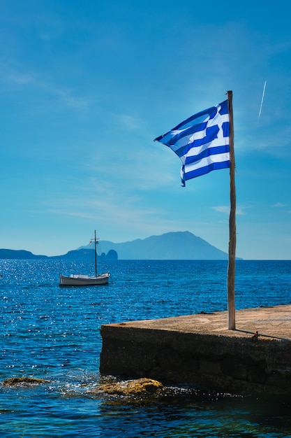 Photo greek flag in the blue sky on pier and traditional greek fishing boat in the aegean sea