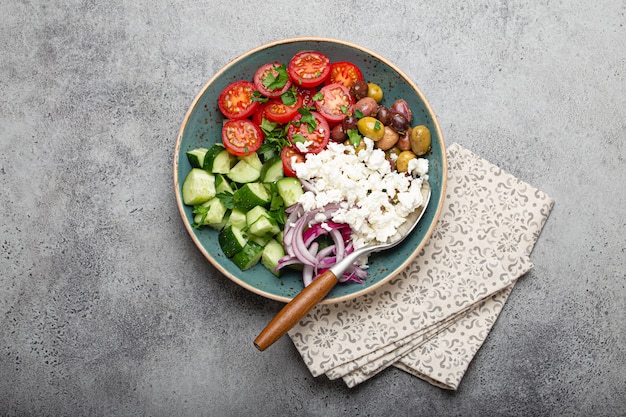 Greek mediterranean salad with tomatoes, feta cheese, cucumber, whole olives and red onion in blue ceramic plate on gray concrete background from above, traditional appetizer of Greece